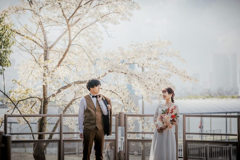 満開の桜の木の下で見つめ合う新郎新婦。お台場海浜公園での春の洋装前撮りの1枚