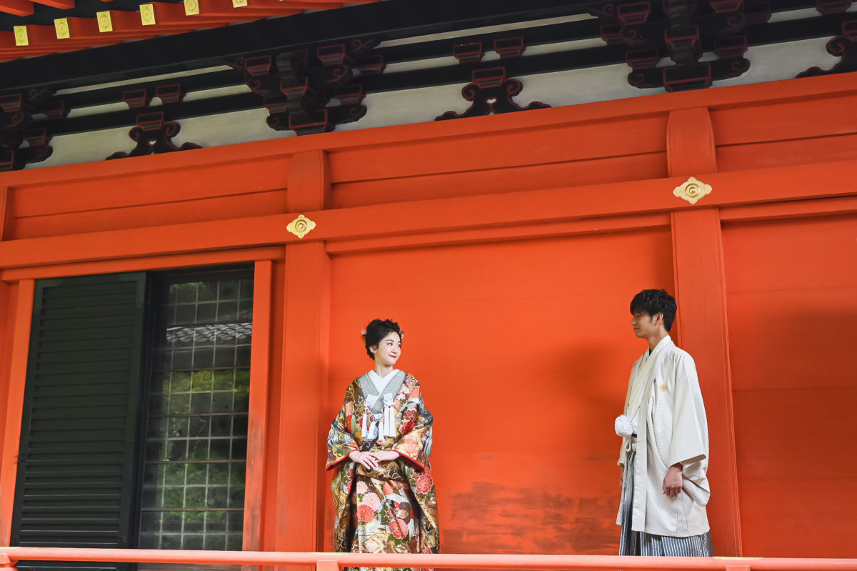 朱色の漆喰壁の前で向き合う和装を着たふたり