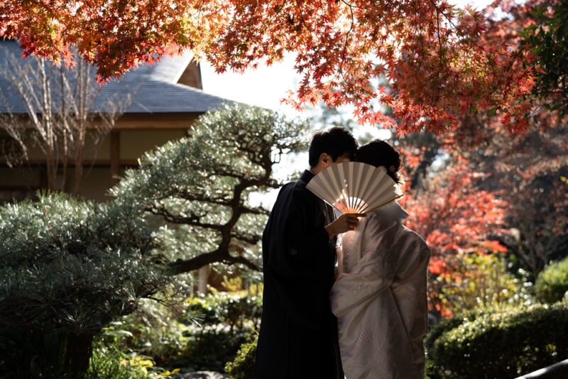 真っ赤な紅葉と松の庭で、扇子で顔を隠して寄り添う紋付袴の新郎と白無垢の花嫁。秋の和装前撮り