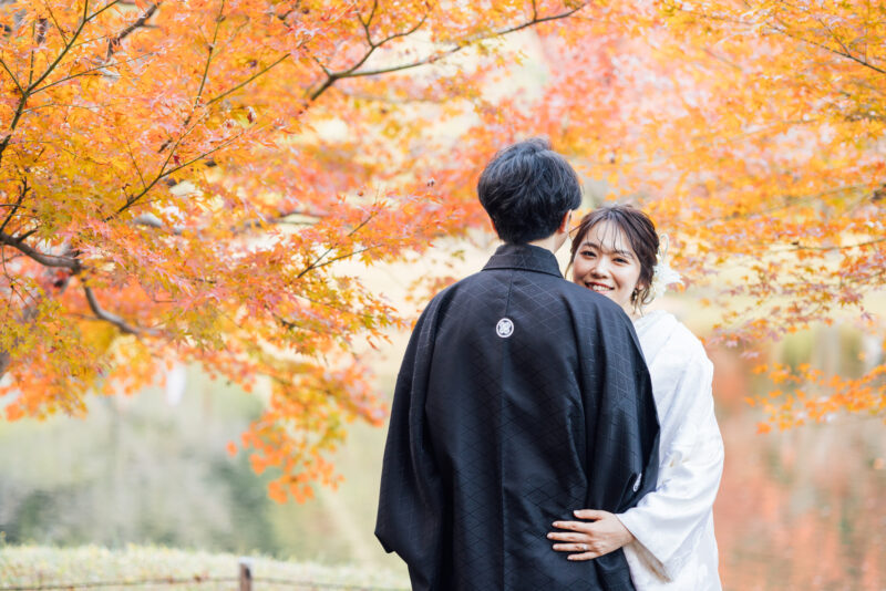 オレンジ色の紅葉に包まれて、新郎に寄り添って微笑む白無垢の花嫁。秋の日本庭園で撮る和装前撮り