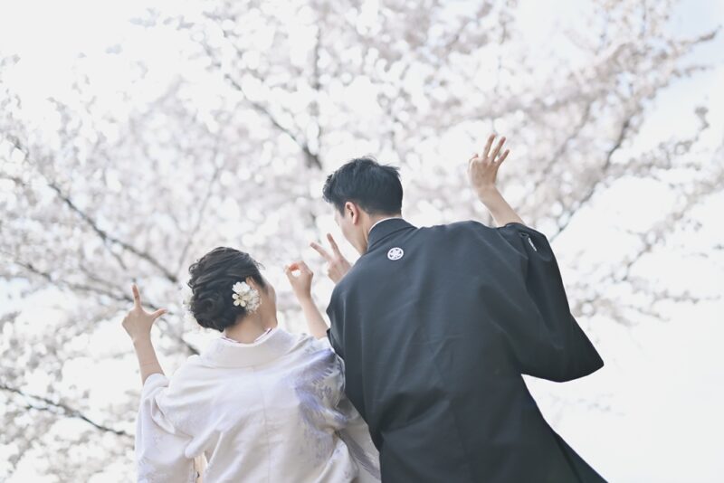 満開の桜を見上げて手を伸ばす和装新郎新婦の後ろ姿