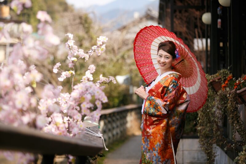 満開の桜の下で朱色の和傘を差す色打掛姿の新婦 古い町並みを背景にした一枚