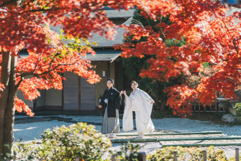 燃えるような紅葉に囲まれた日本家屋の庭を歩く、紋付袴の新郎と白無垢の花嫁。秋の和装前撮り