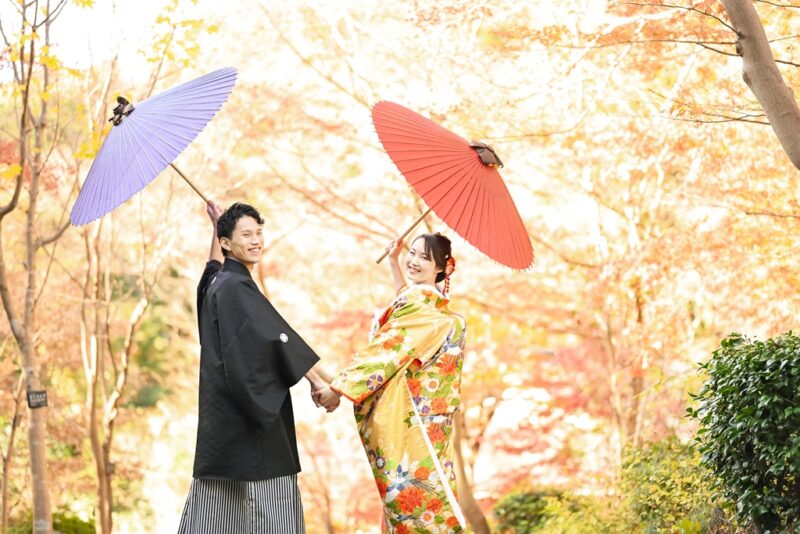 紫と赤の和傘を差して手をつなぐ紋付袴の新郎と色打掛の花嫁。紅葉の日本庭園で撮る和装前撮り