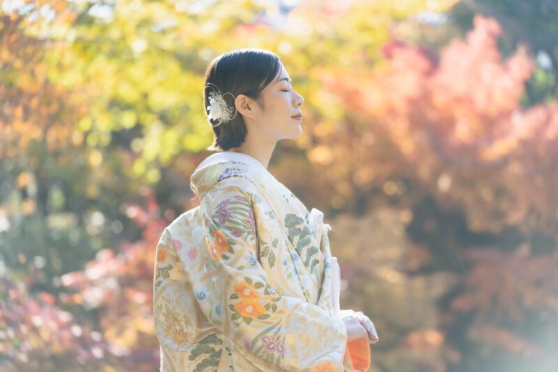 紅葉を背景に目を閉じて陽光を浴びる、白地に花柄の色打掛の花嫁。秋の和装前撮りの花嫁ショット