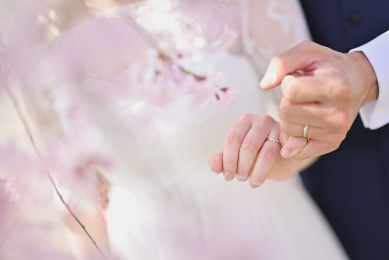 桜の花越しに結婚指輪をつけて手を繋ぐ新郎新婦の手元アップ