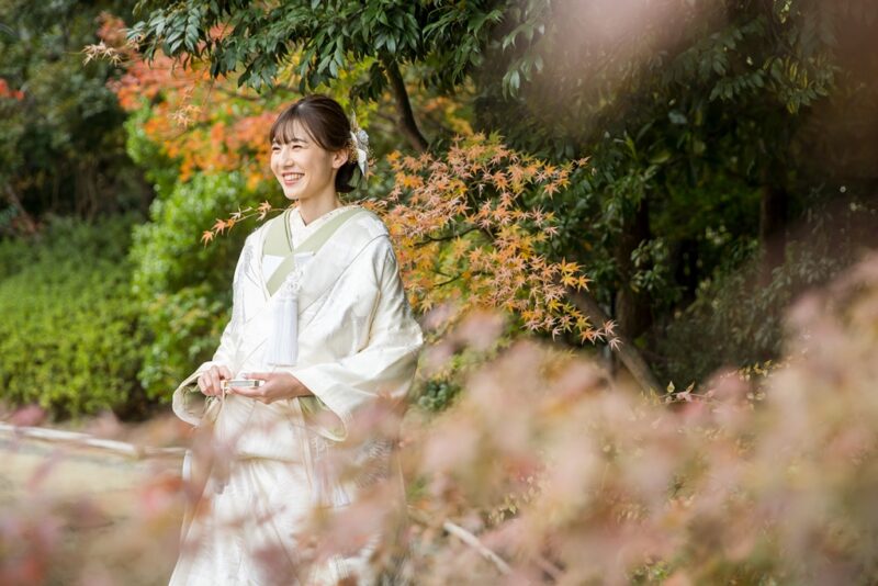 紅葉の前ボケ越しに、やわらかな光の中で微笑む白無垢の花嫁。秋の和装前撮りの花嫁ショット