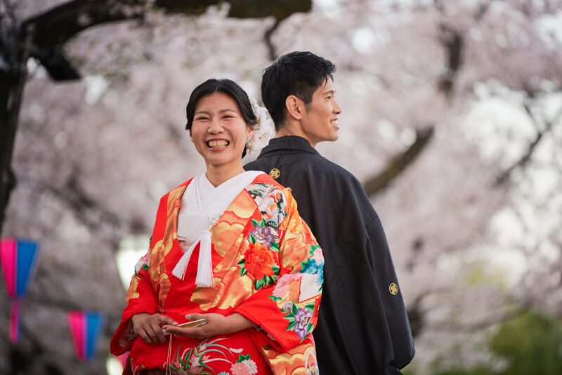 満開の桜を背に笑う赤い色打掛の花嫁と、そばに立つ黒紋付の新郎を写した隅田公園の春の和装前撮り