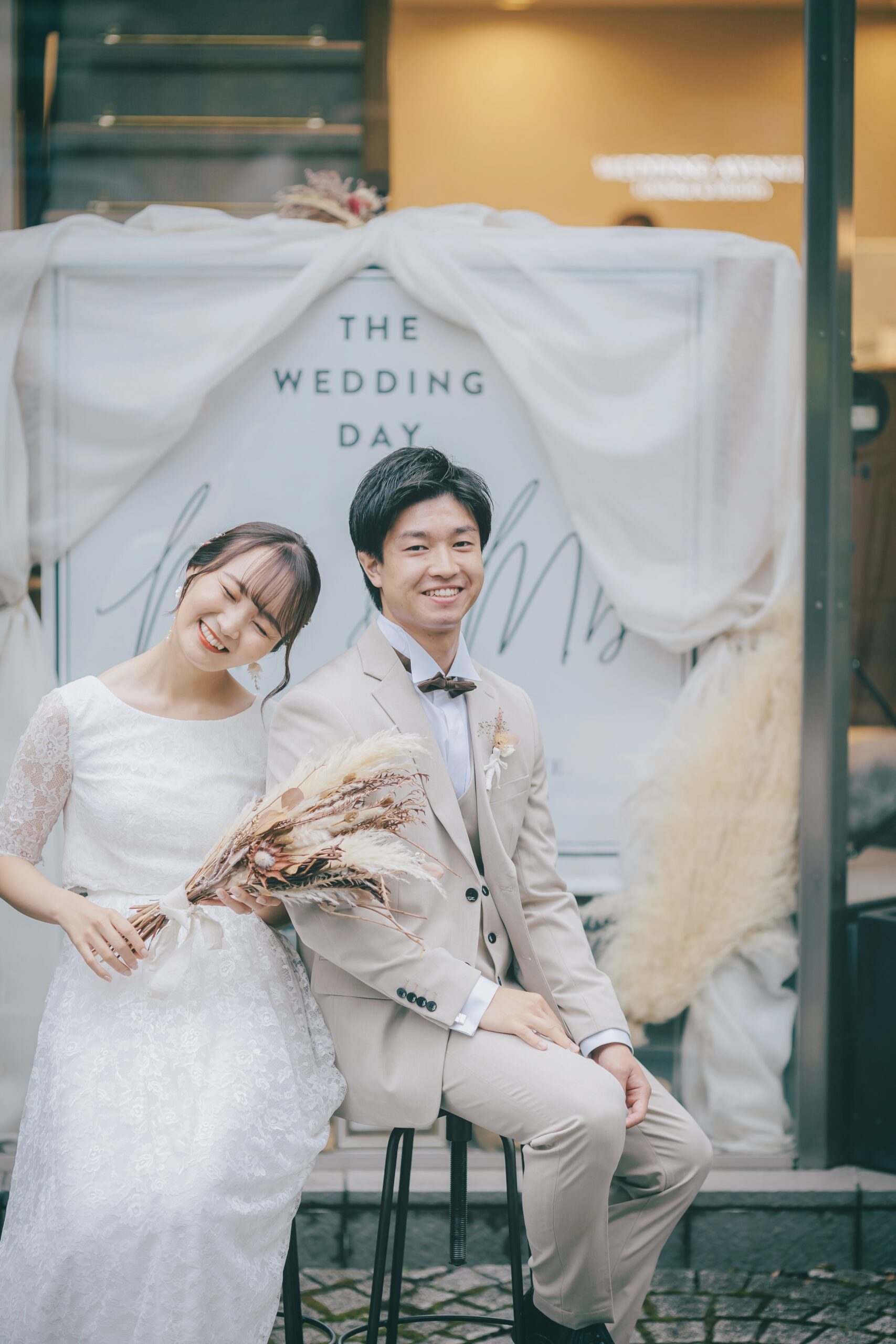 ドライフラワーのブーケを持つレース袖ドレスの新婦とベージュスーツの新郎 店舗前でのショット
