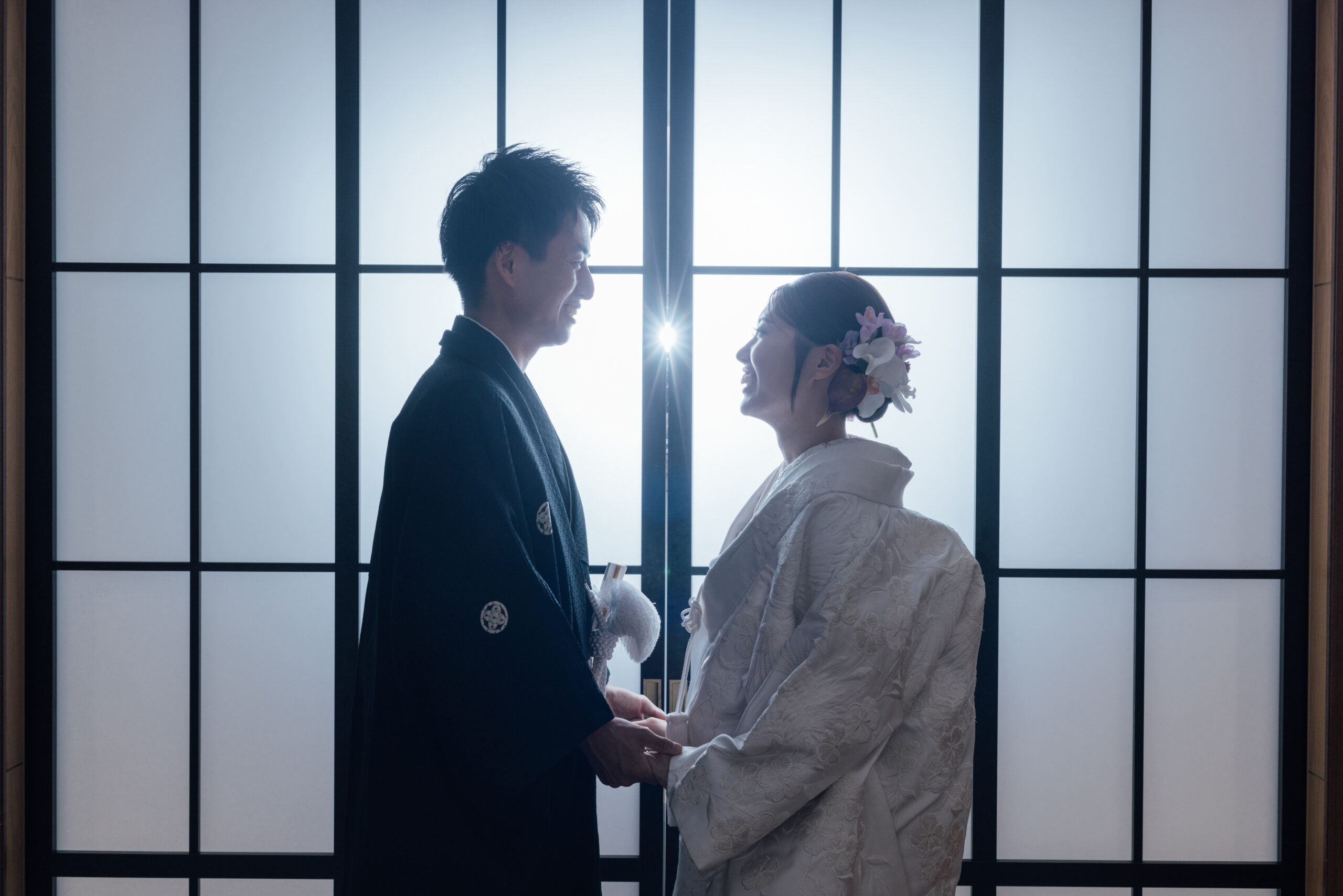障子窓の逆光で見つめ合う白無垢と黒紋付袴の新郎新婦 華やかな髪飾りが映えるシルエット