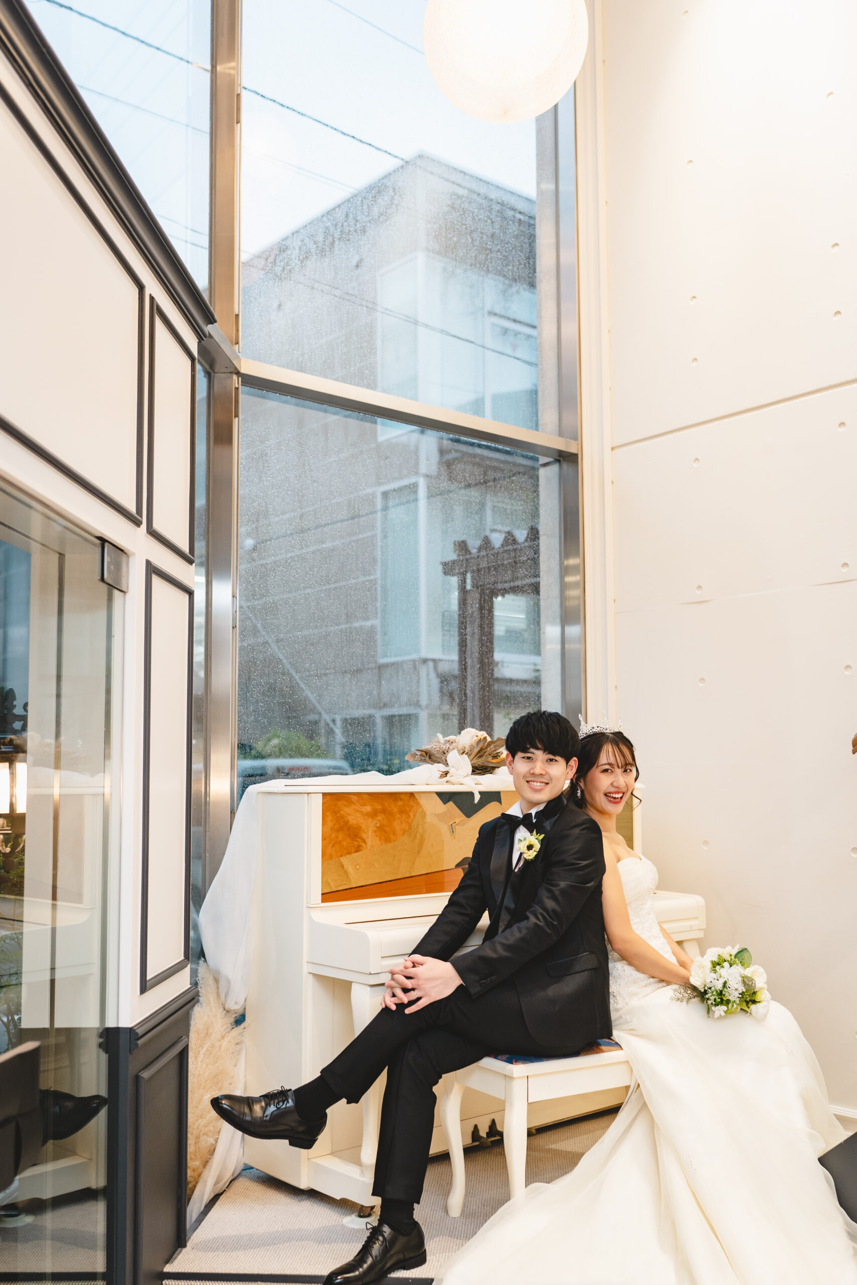 白いピアノに腰掛ける新郎新婦 ティアラを着けたストラップレスドレスと大きな窓が印象的な一枚