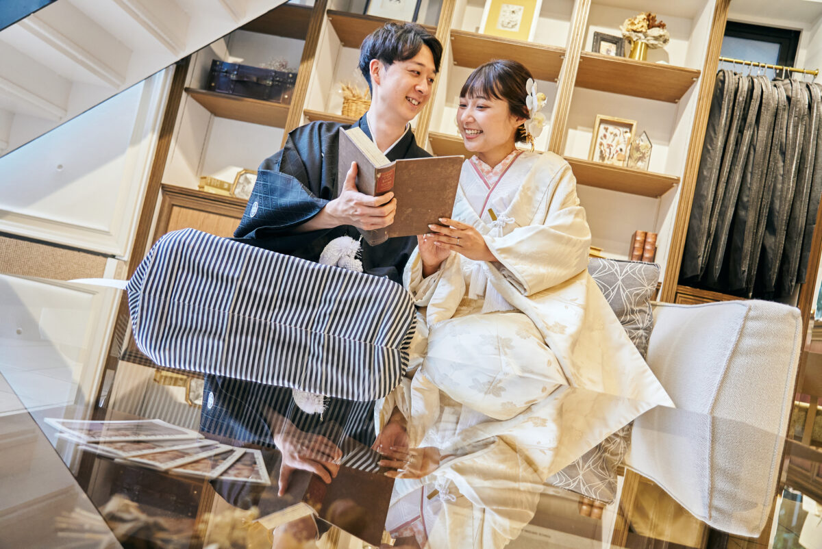 アルバムを一緒に眺めて微笑む新郎新婦 黒紋付袴と生成り色の色打掛 鏡面テーブルの反射が印象的