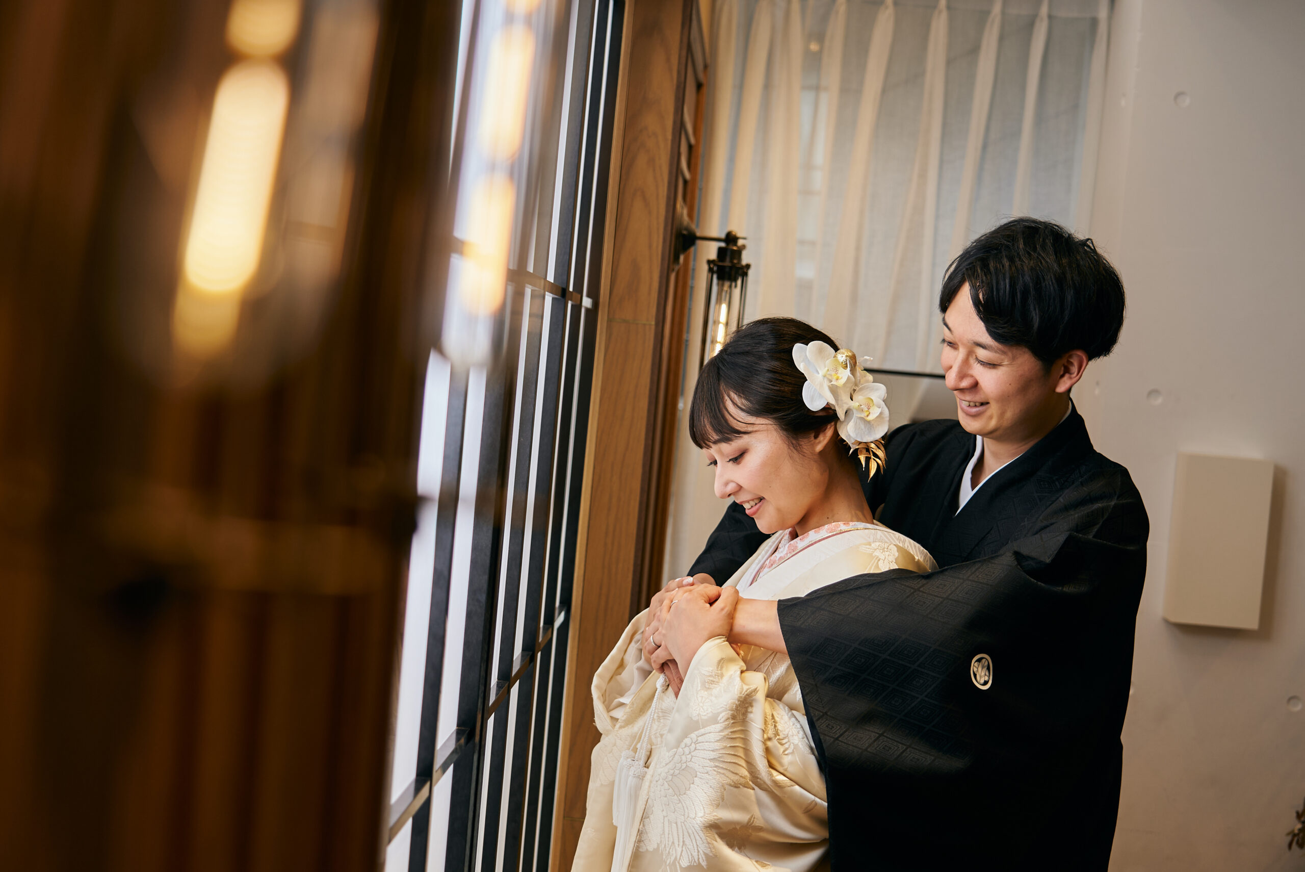黒紋付袴の新郎と金色の色打掛姿の新婦 障子窓辺での温かな触れ合いショット