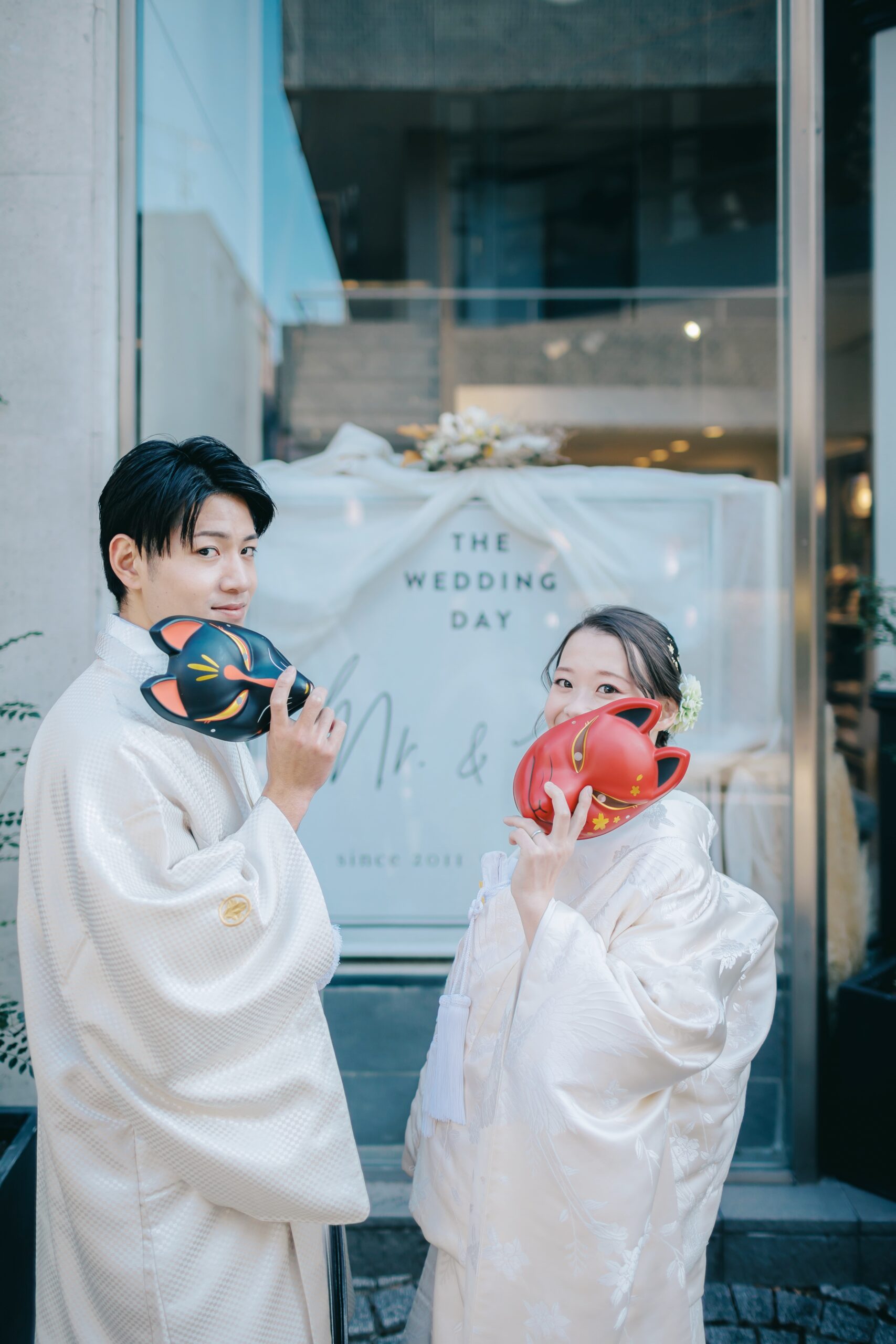 白い和装姿で狐のお面を持つ新郎新婦 店舗入口の「THE WEDDING DAY」ウェルカムボード前
