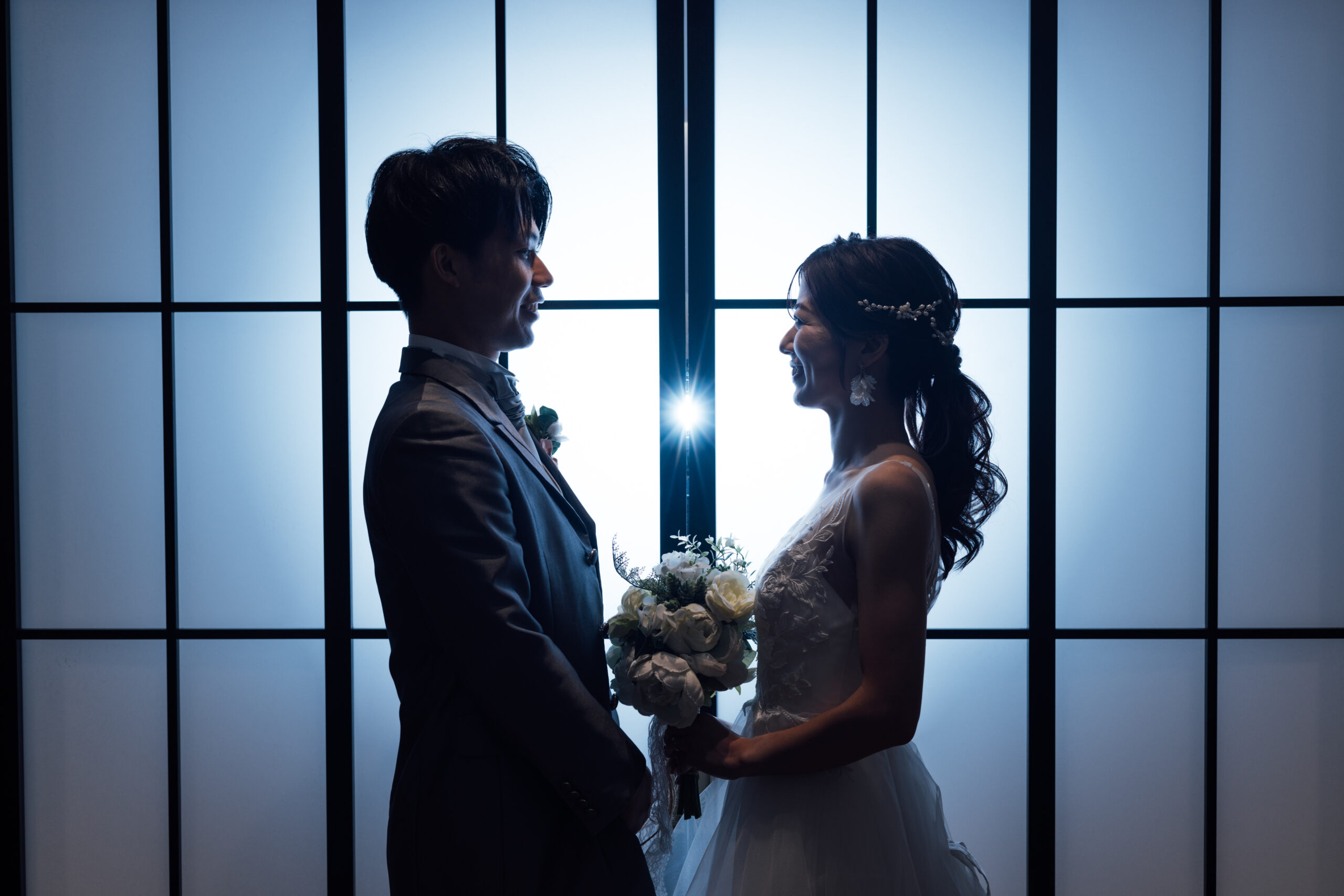 青みがかった逆光の障子窓越しに見つめ合う新郎新婦 ブーケを手にしたシルエットショット