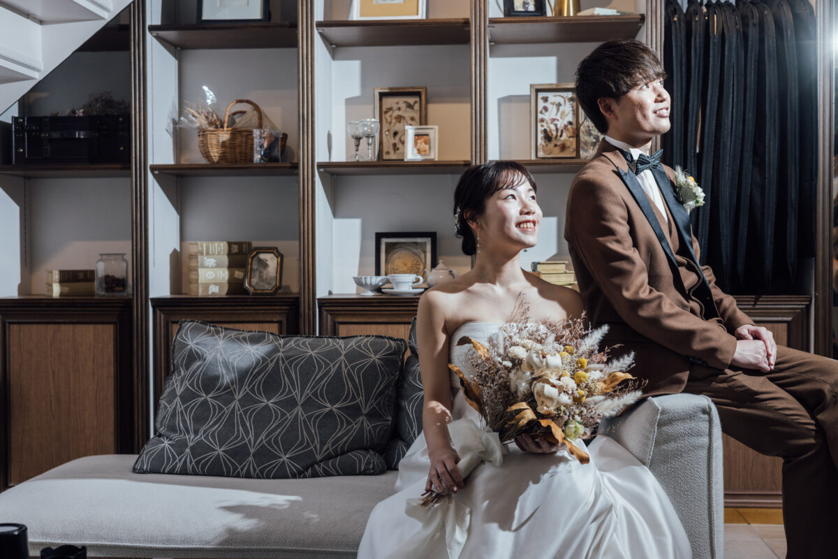 アンティーク書棚を背景にソファでくつろぐ新郎新婦 ブラウンスーツとドライフラワーブーケ