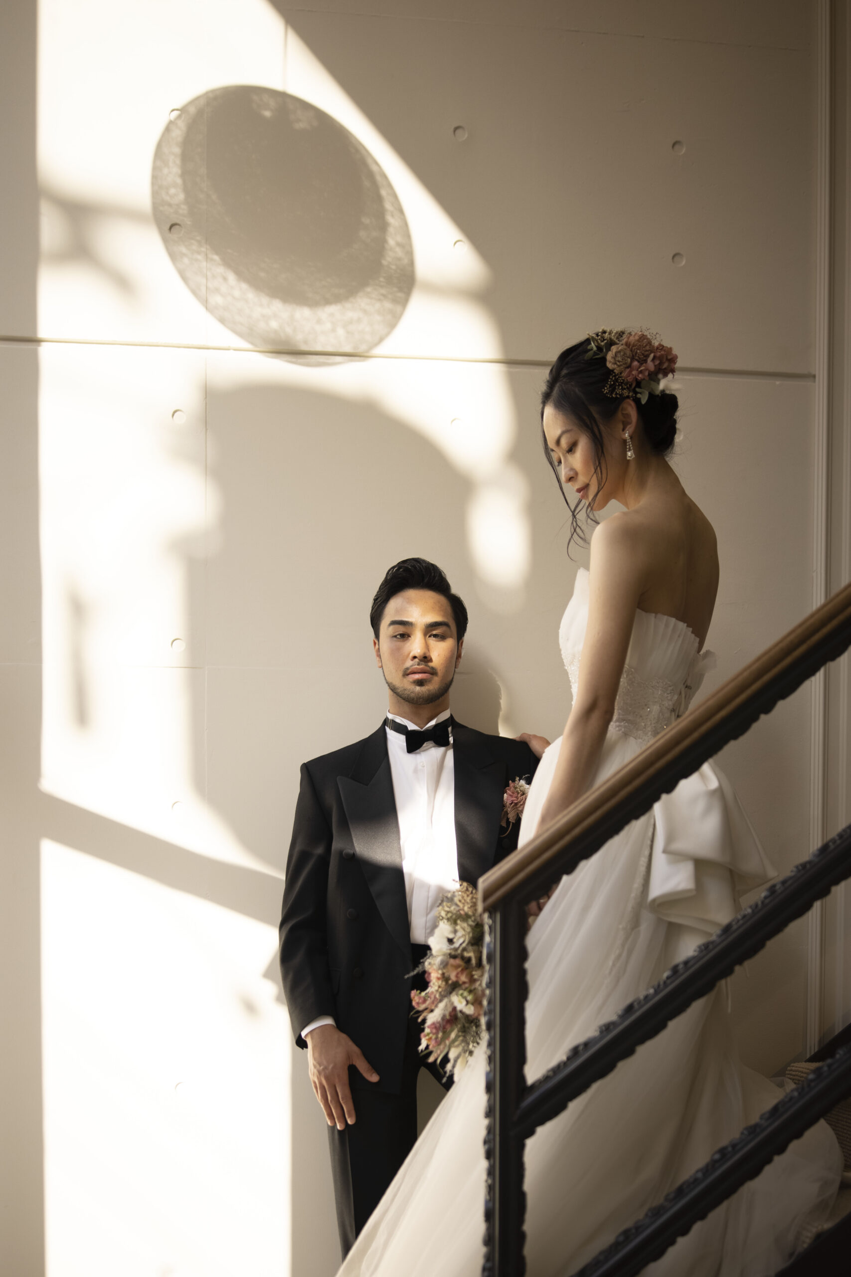 光と影が印象的な階段での新郎新婦 黒タキシードとレースドレス、ドライフラワーブーケ