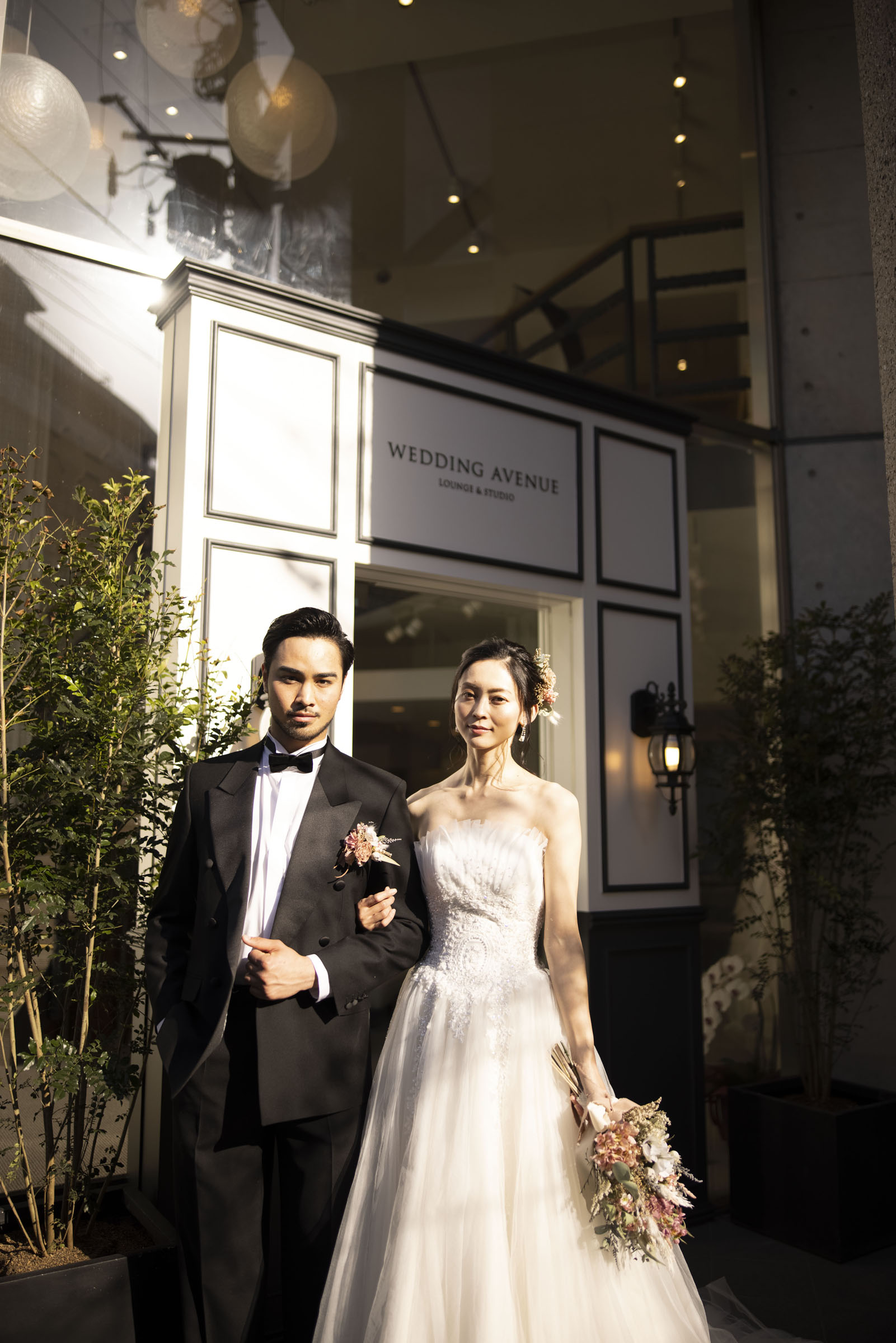 「WEDDING AVENUE」店舗前で寄り添う新郎新婦 総ビジューのストラップレスドレスとドライフラワーブーケ