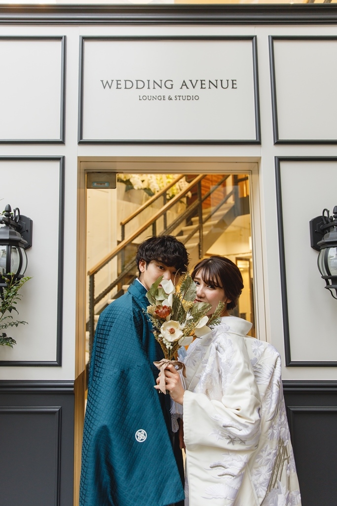 青緑の羽織袴の新郎と白い色打掛の新婦 花束越しに微笑む「WEDDING AVENUE」店舗前ショット