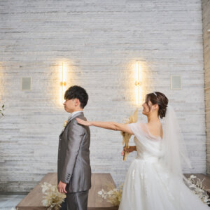 白い石壁の前で背を向けた新郎の肩に手を伸ばす花嫁。チャペルで行うファーストミートの始まりを撮った前撮り写真