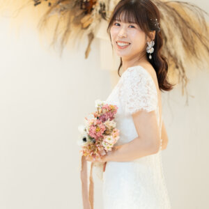 パンパスグラスのスワッグの前で、くすみピンクのブーケを持って振り返るレースのドレスの花嫁。白い背景のナチュラルな前撮り