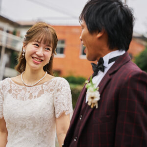 校舎の前で新郎と向き合い、満面の笑みを見せるパールのネックレスの花嫁。ボルドーのタキシードと胸元のブートニアも映える前撮り