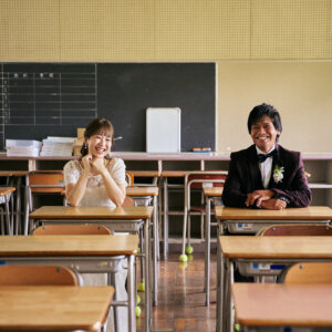 黒板を背に教室の席に座り、こちらを向いてほほえむ新郎新婦。整然と並ぶ机が印象的な学校の前撮り