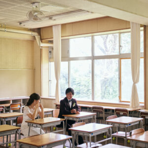 大きな窓のある広い教室で、離れた席に座って笑い合う新郎新婦を引きで写したロケーションフォト