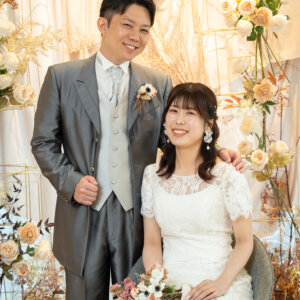 ドライフラワーと白い花のアーチの前で、椅子に座るブーケを持った花嫁と肩に手を添える新郎。クラシカルなスタジオフォト