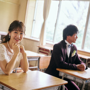 教室の机に両手で頬杖をつき、カメラに笑顔を向ける花嫁と窓の外を見る新郎。学校で撮る洋装前撮りのワンシーン