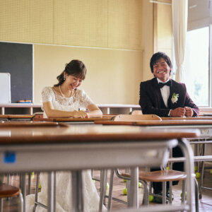 手前の机越しに、教室の席に座ってほほえむ新郎新婦を写したウェディングフォト。懐かしい教室の空気感が伝わる構図