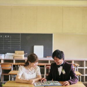 教室の机をくっつけてアルバムをのぞき込み、学生時代を振り返る新郎新婦。思い出の詰まった学校で撮る前撮りのワンシーン