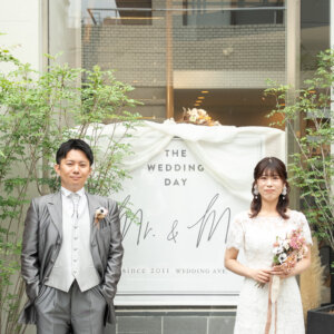 THE WEDDING DAYのボードを飾った南青山の店舗入口で、ブーケを持って並ぶ新郎新婦。グリーンに囲まれた外観で撮る前撮り