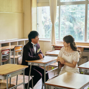 机が並ぶ明るい教室で、隣り合う席に座って向き合う新郎新婦。レースのウェディングドレスとボルドーのタキシードで撮った学校の前撮り