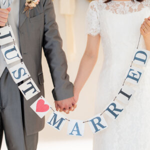 手をつないでJUST MARRIEDのガーランドを掲げる新郎新婦の手元。レースの半袖ドレスがナチュラルなスタジオフォト