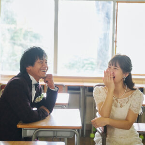 窓際の席に座り、顔を見合わせて笑い合う新郎新婦。光がたっぷり入る教室で撮ったナチュラルな結婚写真