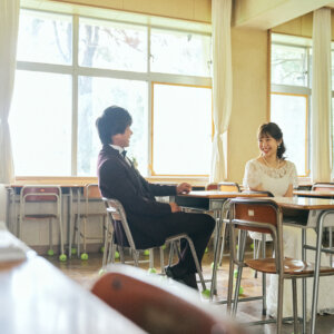 手前の机越しに、教室の席で向かい合って話す新郎新婦を写した一枚。学校ロケーションフォトらしい奥行きのある構図