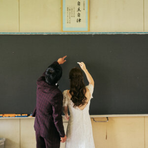 黒板の前で並んで腕を伸ばし、チョークで絵を描く新郎新婦の後ろ姿。教室で撮る洋装前撮りのワンシーン