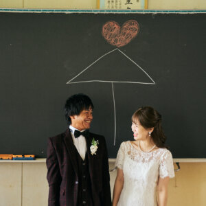 黒板に描いた相合い傘とハートの下で、見つめ合って笑う新郎新婦。学校の前撮りで人気の黒板アートのポーズ