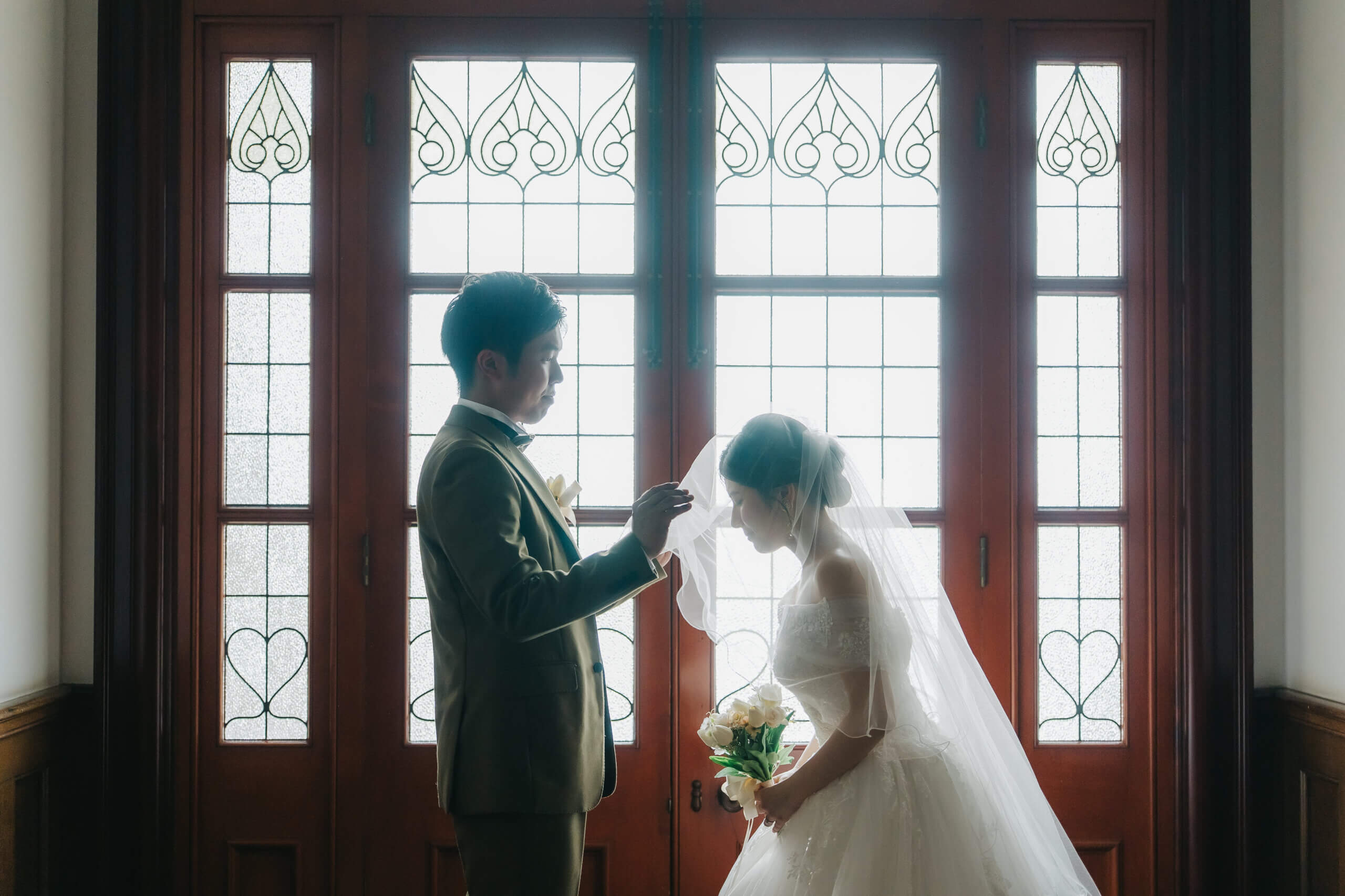 旧古河邸のステンドグラスの扉の前で、ブーケを手に向き合うタキシードの新郎とウェディングドレスの新婦の洋装前撮り