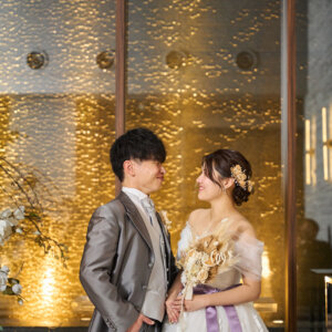 金色の壁が輝くチャペルで、ラベンダーのサッシュのウェディングドレスの花嫁とグレーのタキシードの新郎が見つめ合う前撮り写真