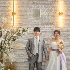 ドライフラワーのスワッグが飾られた白い石壁の前に、手をつないで並ぶ新郎新婦。チャペルで撮る洋装フォトウェディングの全身ショット