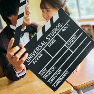 カチンコを手にした新郎と、その後ろで笑う花嫁。机が並ぶ教室で撮った映画のワンシーンのような前撮り
