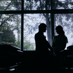 薄暗い教室の窓辺で向かい合う新郎新婦のシルエット。窓の外の緑が映える映画風のウェディングフォト