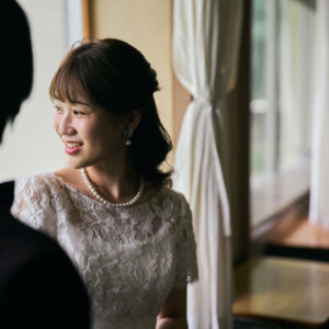 教室で新郎越しにほほえむパールのネックレスとレースのドレスの花嫁。学校の前撮りで撮った自然な表情