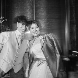 新郎のジャケットを羽織った花嫁と、ベスト姿の新郎がほほえみ合うモノクロの前撮り写真。チャペルの装花の前で動きのあるブレが雰囲気を添える