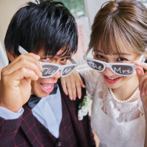 'The Mr''The Mrs'と書かれた白いサングラスを指でずらしてのぞき込む新郎新婦。小物で遊ぶ学校の前撮りのワンシーン