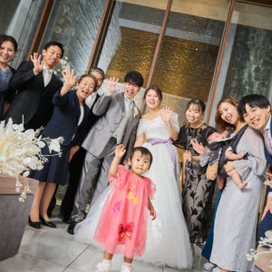 チャペルの祭壇前で、家族や友人と一緒に笑顔で手を振る新郎新婦とピンクのワンピースの娘。にぎやかな家族写真を残すフォトウェディング