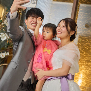 チャペルの金色に輝く壁の前で、小さな娘を抱いてスマホで自撮りする新郎新婦。家族3人で残すあたたかなチャペルフォト