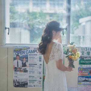 窓辺でひまわりのブーケを抱え、背中の開いたレースのドレスで外を見つめる花嫁の後ろ姿。学校の教室で撮る洋装前撮り